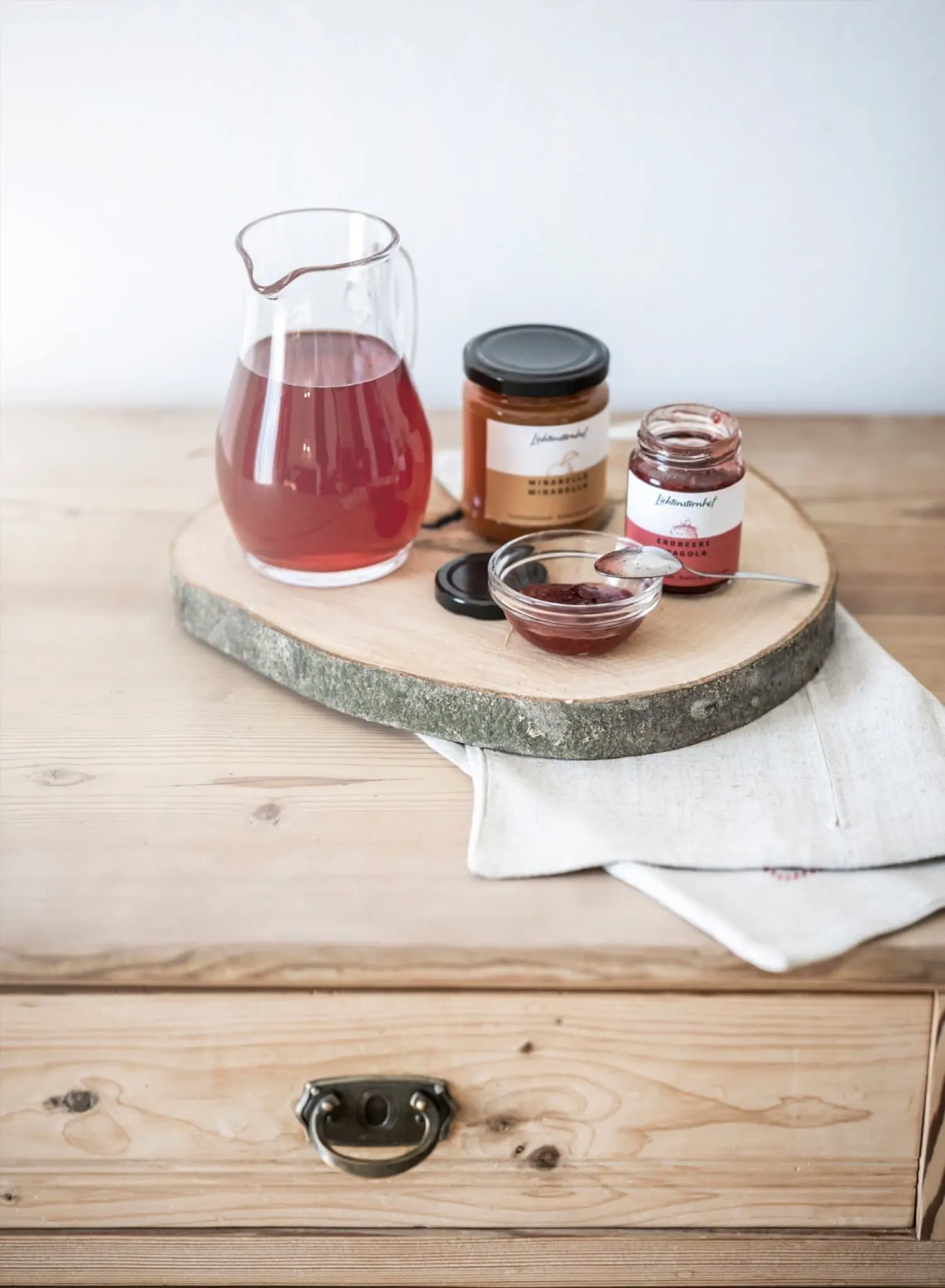 Glaskaraffe mit rotem Sirup und zwei Gläsern Fruchtaufstrich auf einer Baumscheibe, daneben ein Leinentuch