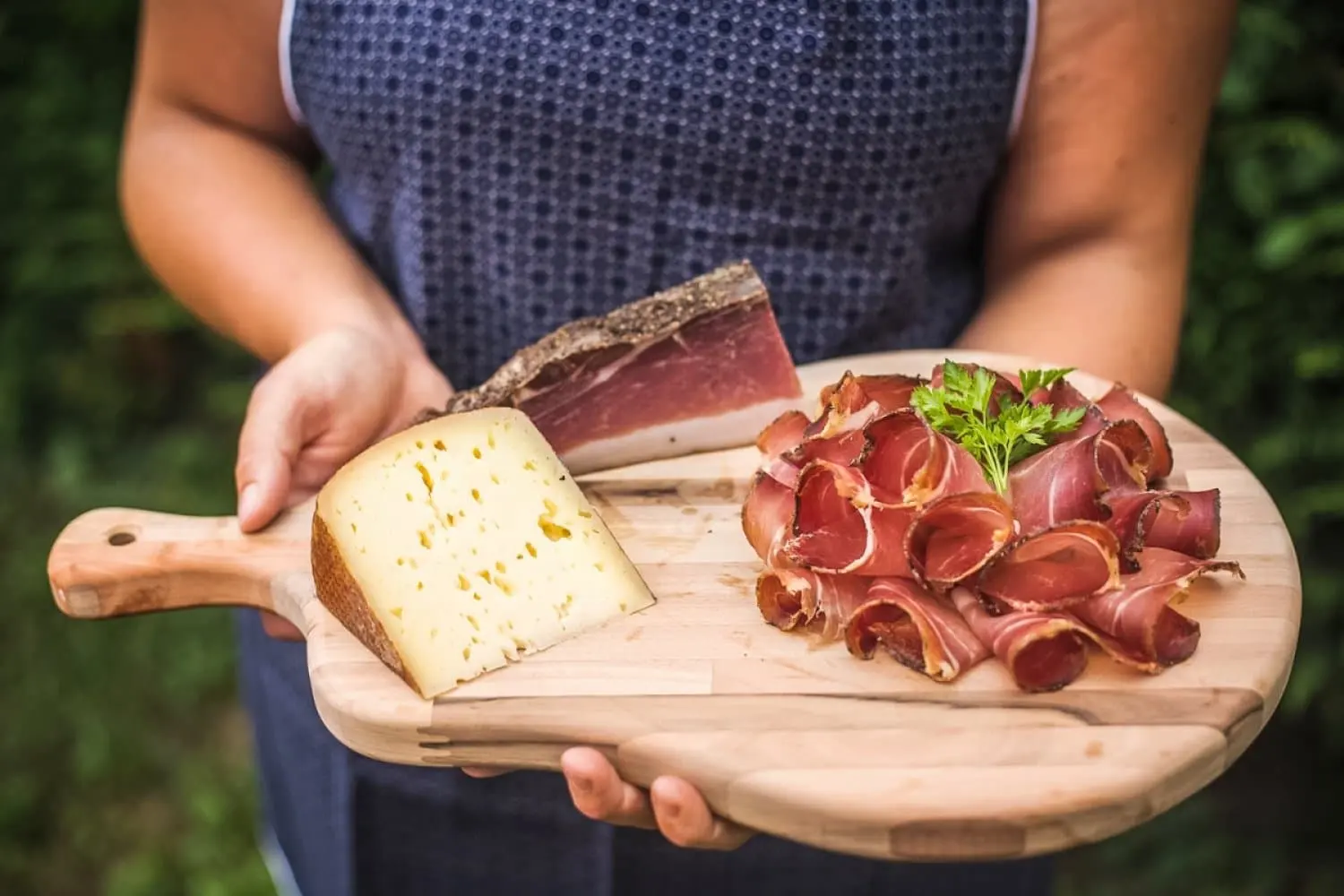 Bäuerin hält ein Holzbrett mit Käsestück, Speckseite und aufgerolltem Speck in die Kamera
